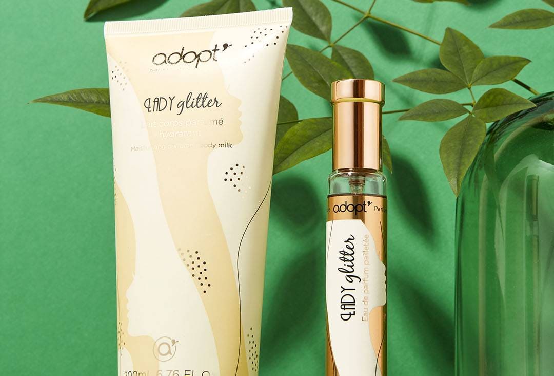 Gros plan sur le lait corporel et le parfum en spray de la marque « adopt », avec des feuilles vertes en arrière-plan, mettant en valeur les emballages beiges et dorés des produits de soin et des parfums.