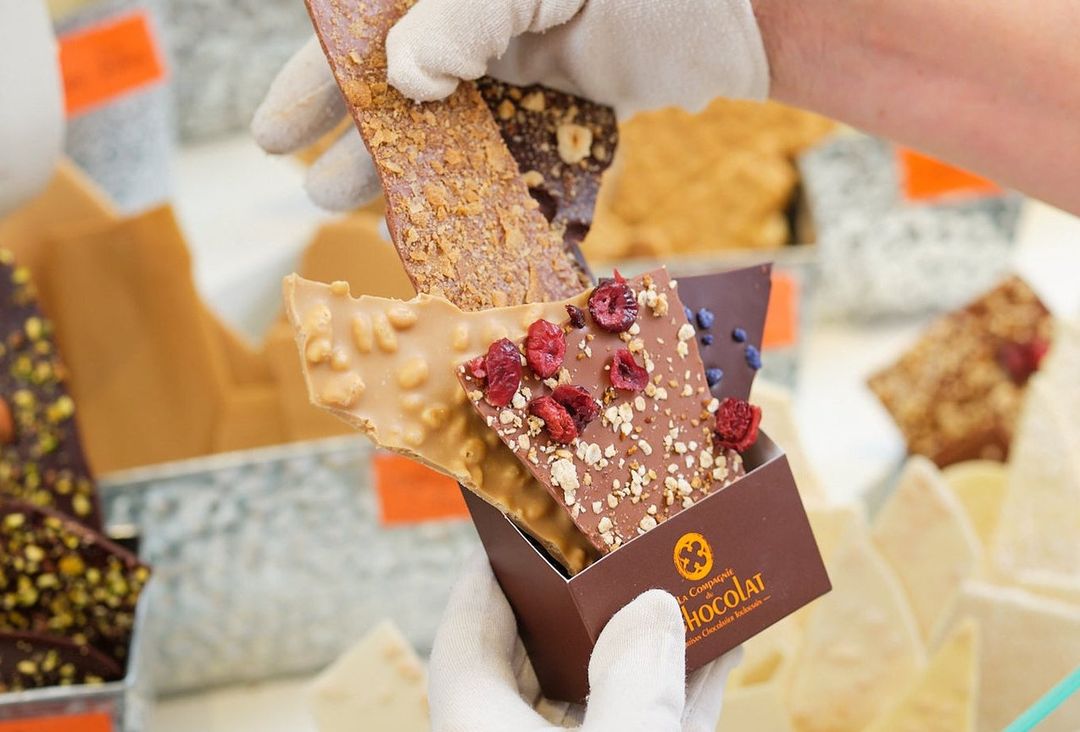Mains gantées présentant des tablettes de chocolat artisanales aux fruits secs et fruits rouges, au-dessus d’un coffret La Compagnie du Chocolat
