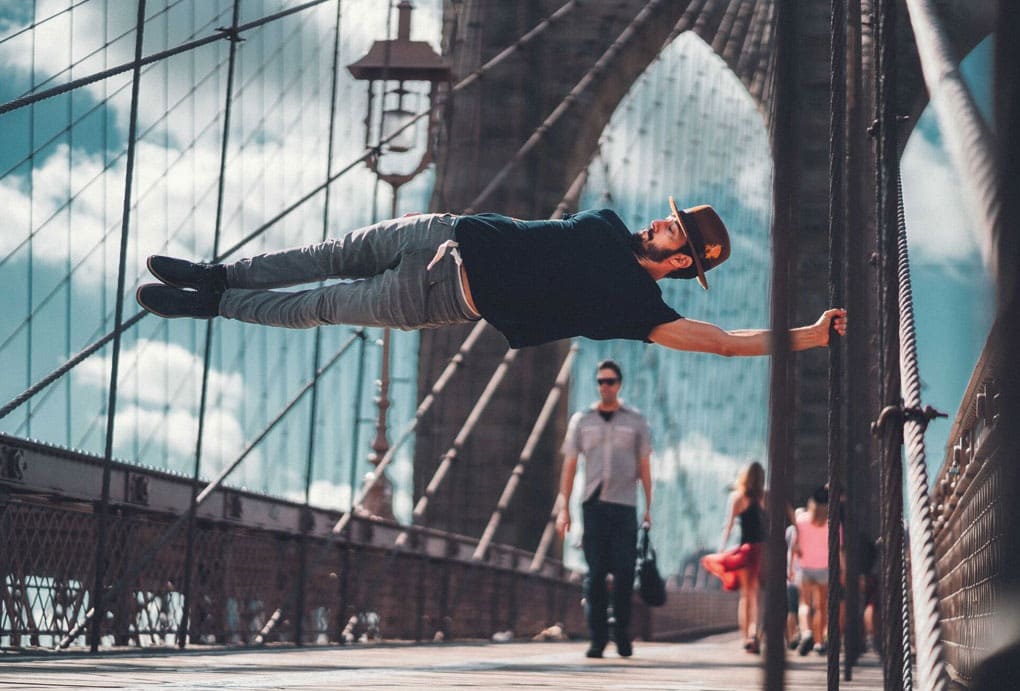 Homme suspendu horizontalement à un câble du pont de Brooklyn, chapeau et t-shirt Pullin